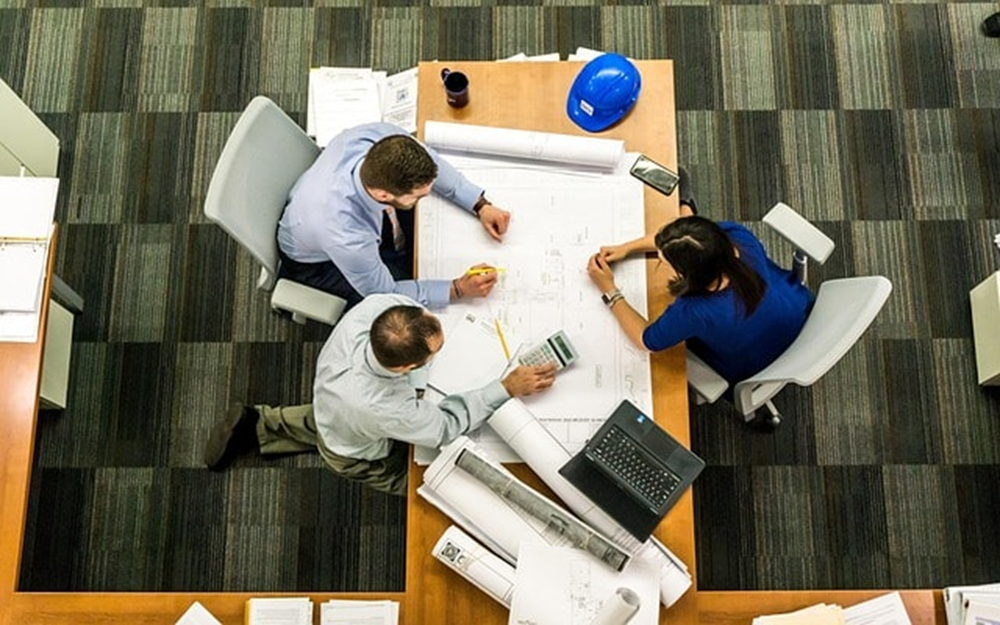 Top down view of three people sitting at a table reviewing documents and plans.