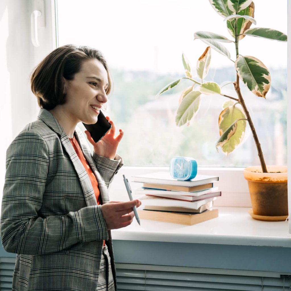 Woman on phone near window with plant, books.